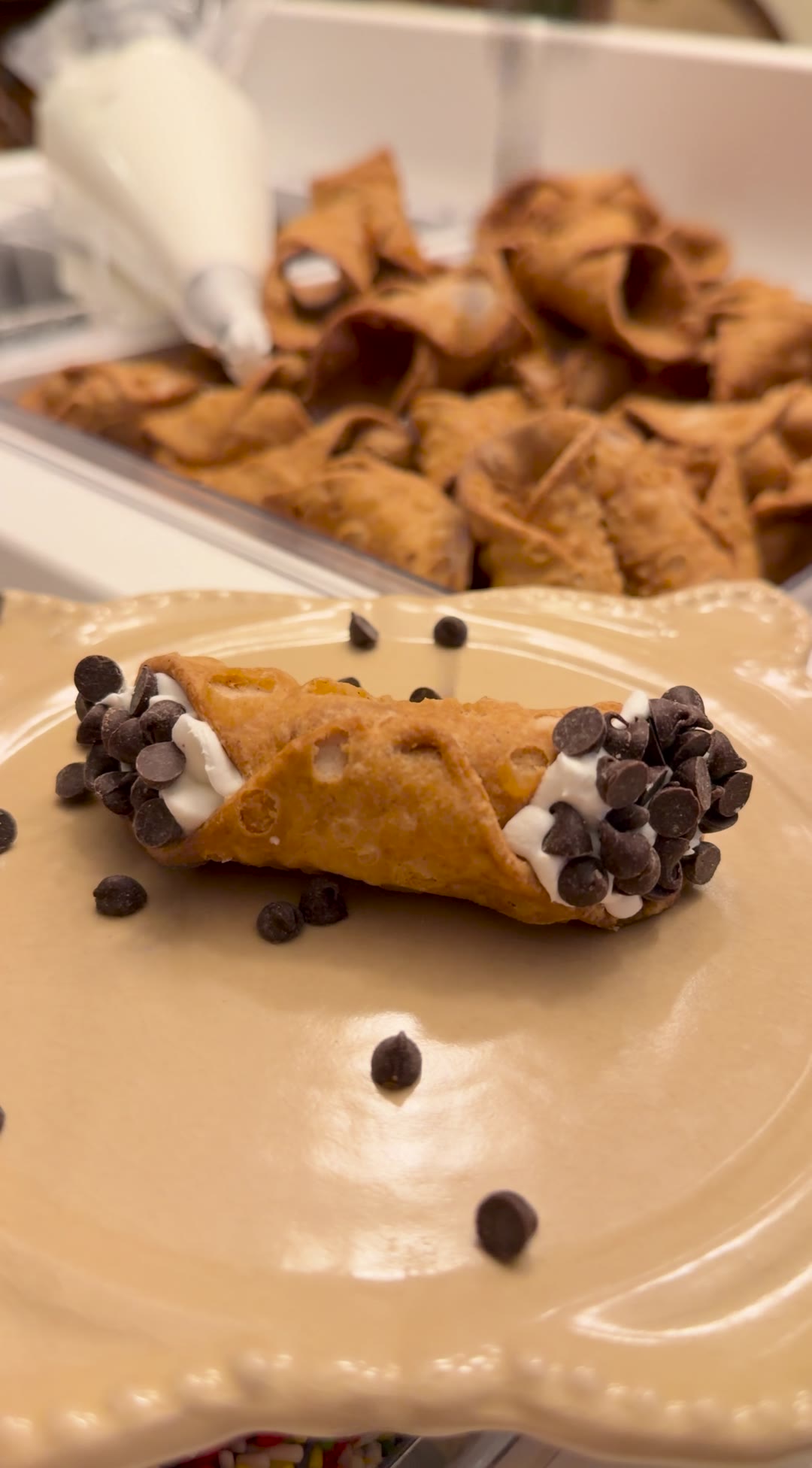 A freshly filled cannoli finished with chocolate chips, with a tray of shells behind